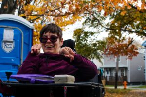 Vicky Lehack在闲置公园旁边的人行道上微笑。她坐在她的轮椅上，紫色毛衣和深色太阳镜。