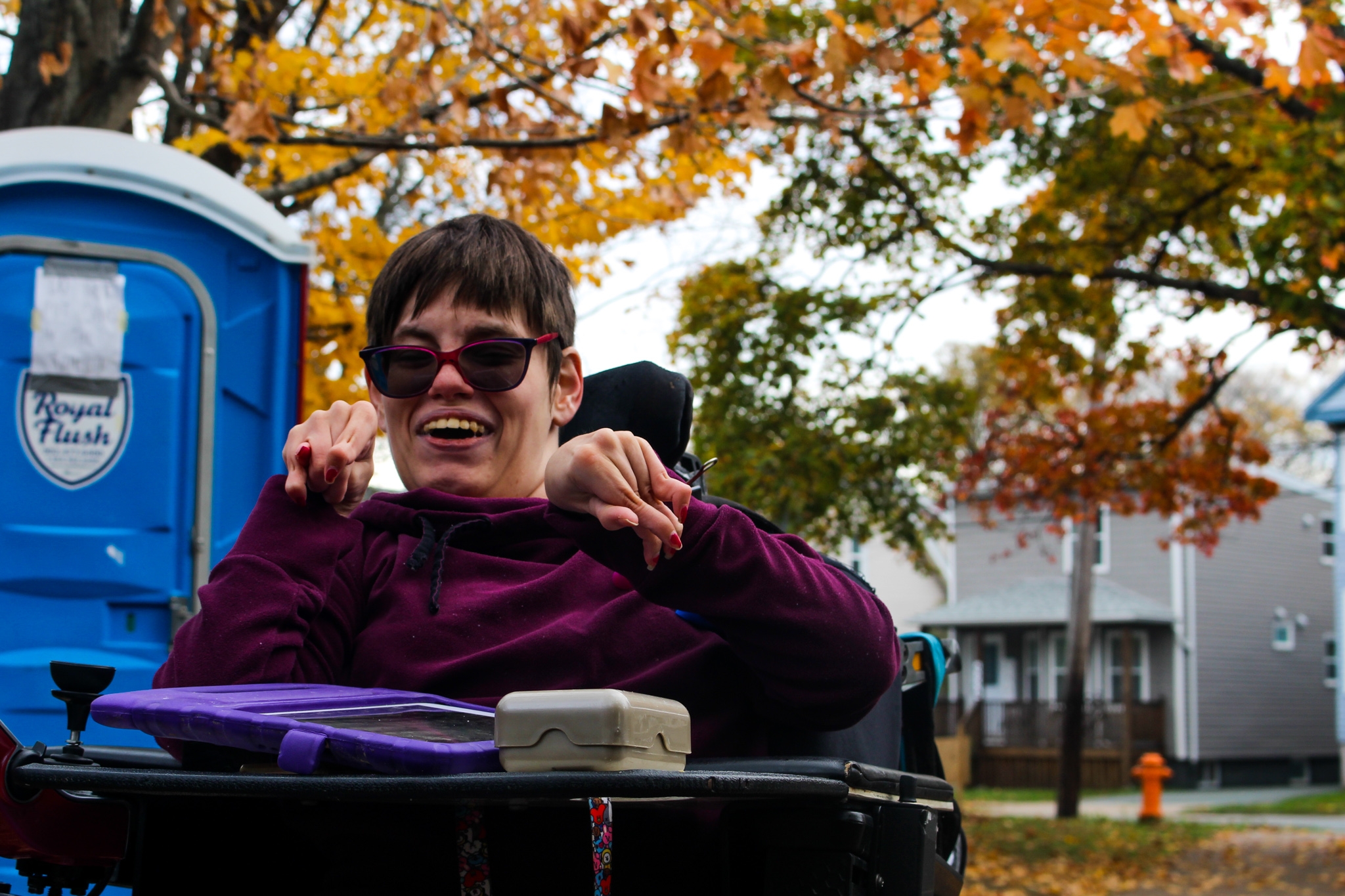 维姬·莱瓦克在米格尔公园旁边的人行道上微笑。她穿着紫色毛衣，戴着深色墨镜，坐在轮椅上。