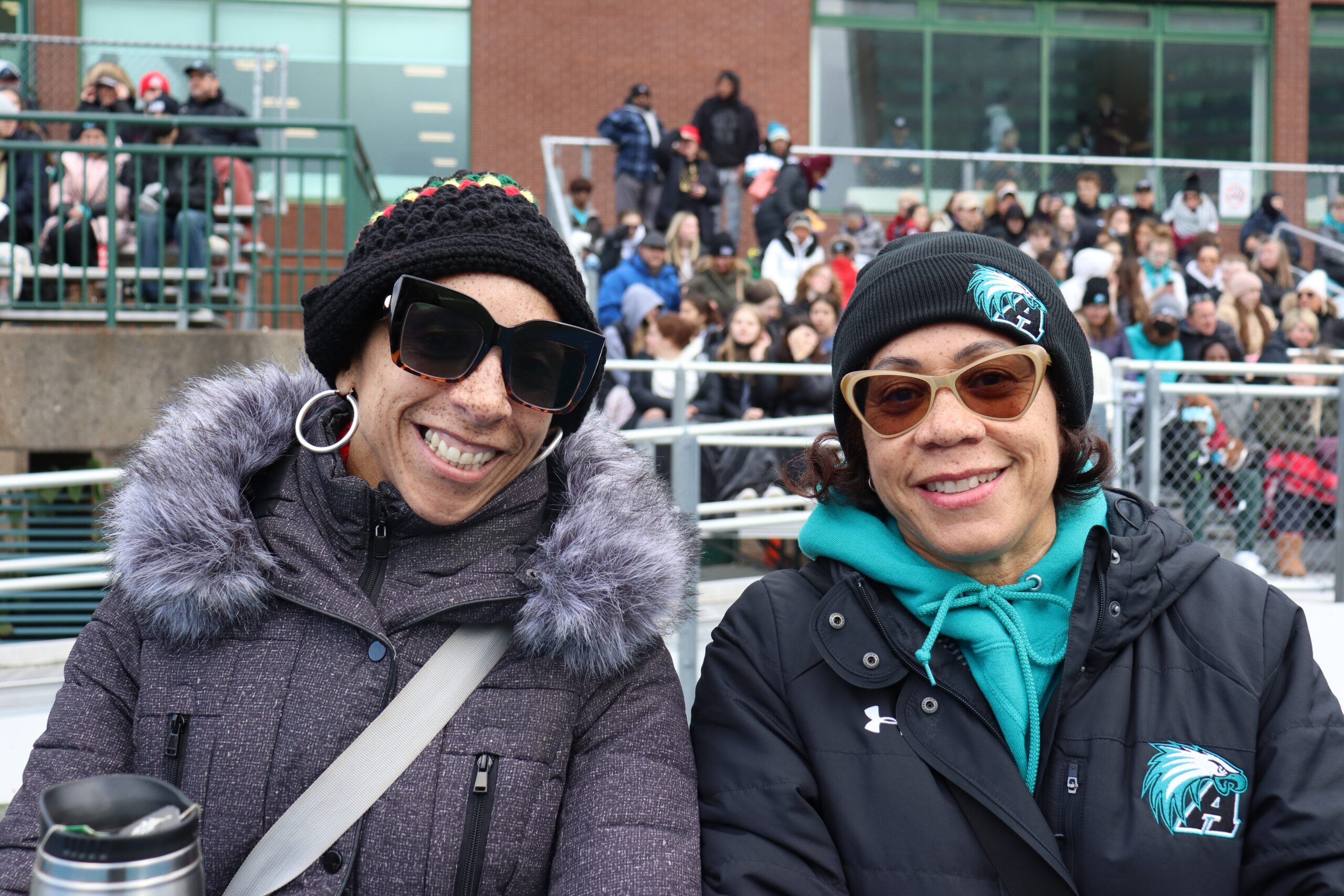 黑人女性玛莎·哈德逊·阿什和卡伦·哈德逊对着镜头微笑，背景是坐在看台上的高中橄榄球迷。两位女士都穿着保暖的皮大衣、帽子和太阳镜。
