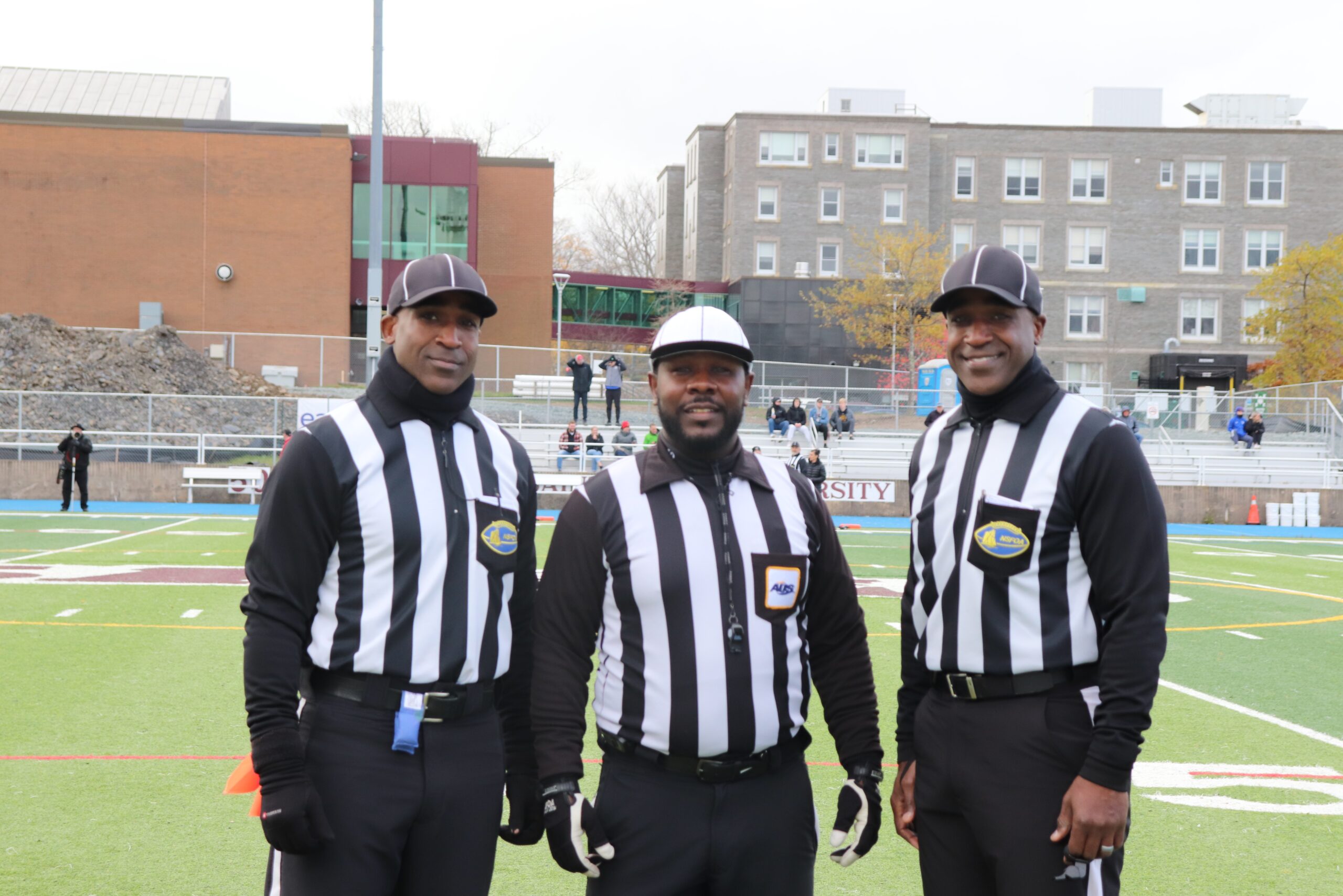 三名黑人足球裁判对着镜头微笑。他们穿着黑白条纹衬衫，戴着棒球帽。他们站在圣玛丽大学的足球场上。