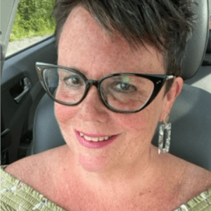 乔安妮·伯纳德是一位迷人的白人女性，棕色短发，戴着时髦的黑框眼镜。在一张自拍中，她微笑着，在她的车里，穿着一件可爱的春天绿色的漂亮上衣。