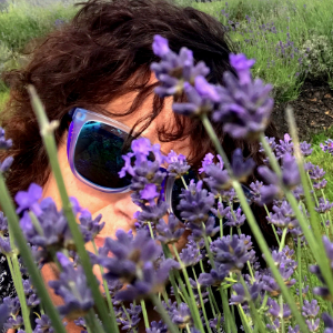 乔迪·德隆，一个留着浓密红褐色头发、戴着大墨镜的白人女人。她正从一片绿色田野里的野生薰衣草后面向外张望。这味道可能很好闻…