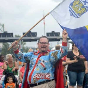 Michael McCluskey, a white man with short dark hair, goatee, and funky glasses. He's shown here in a parade, smiling happily in a Battlestar Galactica shirt and matching cape, carrying a Nova Scotia flag. The shirt and cape fabric have a blue background with comic-style illustrations of space ships and Cylons, and bright red cuffs and collar, and the cape is lined in red.