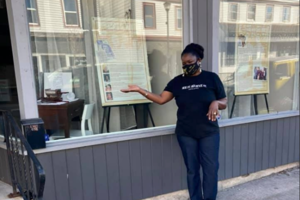 戴着面具、穿着黑色t恤、戴着深色帽子的黑人妇女萨曼莎·迪克森·斯劳特站在人行道上，指着商店橱窗里陈列的展品。