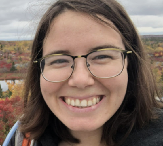 戴着眼镜的年轻白人女子微笑着，直视镜头。她在一个看起来像是明媚秋日的户外。