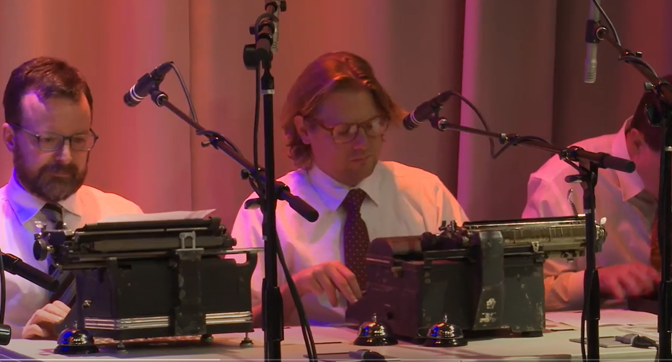 三个穿着白衬衫、打着领带的中年白人男子并排坐在一张桌子旁。每个人面前都有一台打字机，每台打字机上方都悬挂着一个麦克风。最右边的人只隐约可见，他的头低在打字机上。