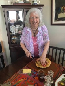 用户莎伦·弗雷泽站在餐桌旁切佛卡夏面包的照片。沙伦身后是一个角落的厨柜，里面陈列着精美的菜肴。莎伦在笑，因为佛卡夏面包很好吃。