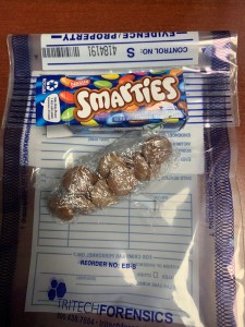 一张小包装Smarties的照片，显示Smarties失去了美味的糖果涂层。现在所有的棕色聪明豆都用保鲜膜包裹着，而聪明豆盒子则放在糖果上面。这张照片是由肯特维尔警察局拍摄的，所以糖果盒和用保鲜膜包装的糖果都在一个证据袋里。