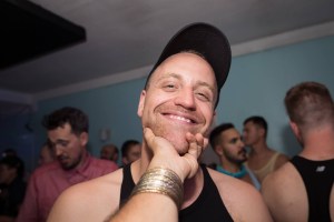 这张照片里，斯图尔特·莱杰雷对着镜头微笑，摄影师伸出手去捏他的下巴。Legere在一个聚会上，其他人在后面。Legere戴着一顶黑色棒球帽和黑色背心。