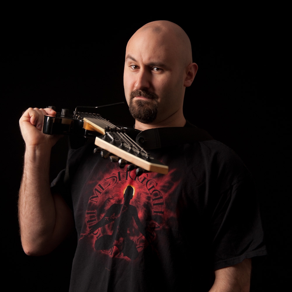 一个留着胡子的秃顶白人男子穿着一件黑色重金属乐队t恤，上面模糊地写着字。他身体前倾，握着吉他颈，使枪托指向观众。