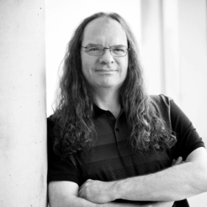 A black and white photo of Tim Covell, a white man with long wavy brown hair and wire-rimmed glasses. He's wearing a dark short-sleeved shirt, leaning against a wall with his arms crossed.