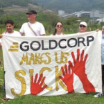 人们举着一条织物横幅，上面写着“黄金公司让我们生病”，上面画着巨大的红手。