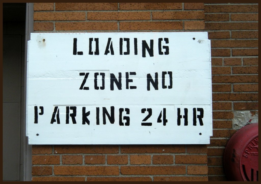 砖墙上的模板标志。上面写着装货区24小时禁止停车。这些字母都是大写的，有些是歪歪扭扭的，线条断掉的样子看起来就像这个地方叫
