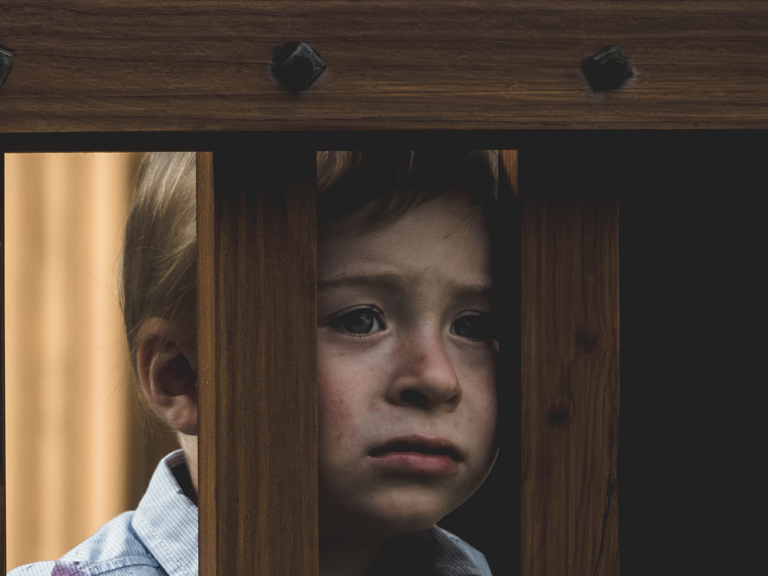 一个年轻的，看起来很悲伤的孩子透过一个木制的操场设备甲板的板条向外张望。