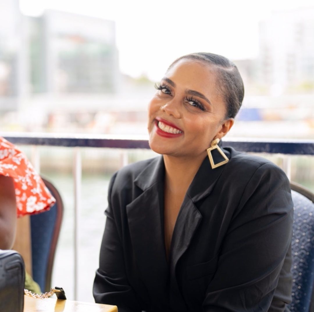 A photo of René Boudreau smiling for the camera. She's wearing a black suit and has large square golden earrings. Her hair is slicked back with a side part and she has on red lipstick.