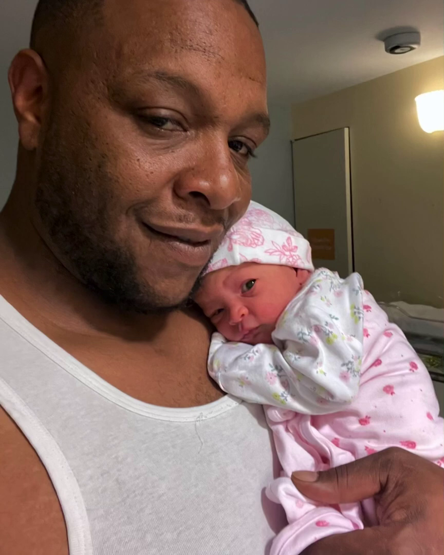 照片中的亚历克斯·托马斯抱着一个婴儿，婴儿穿着粉色和白色相间的卧铺，还裹着粉色图案的毯子。