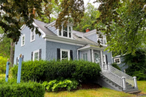 在绿色的树木和灌木丛中，映衬着一幢白色装饰的灰色房子。房子前面有一段长长的楼梯，二楼有两个天窗向前延伸。这张照片是从房子的左侧拍摄的。