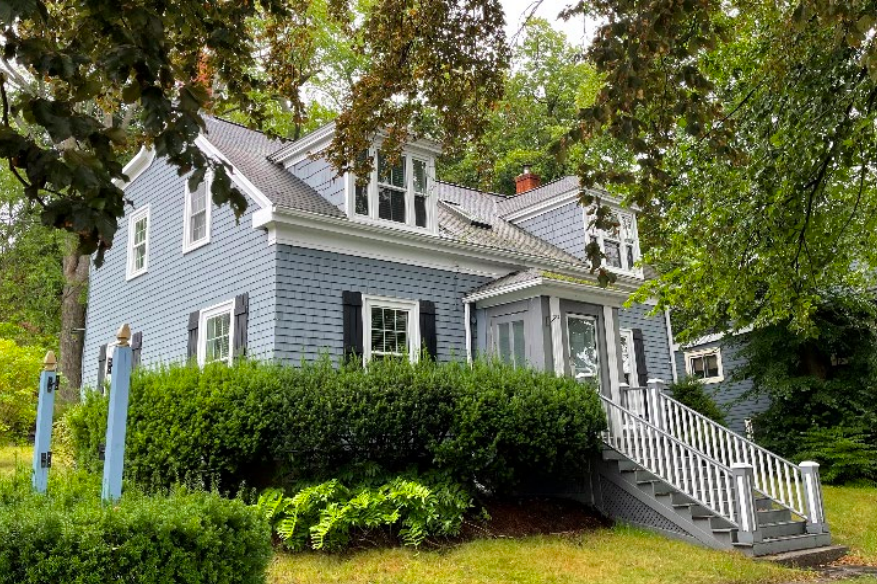 灰色的房子有白色饰边的灰色房子在绿树和灌木丛中被出现。该房子在前面有一个长长的楼梯，两个休眠者伸出第二级。照片从家里​​的左侧拍摄。