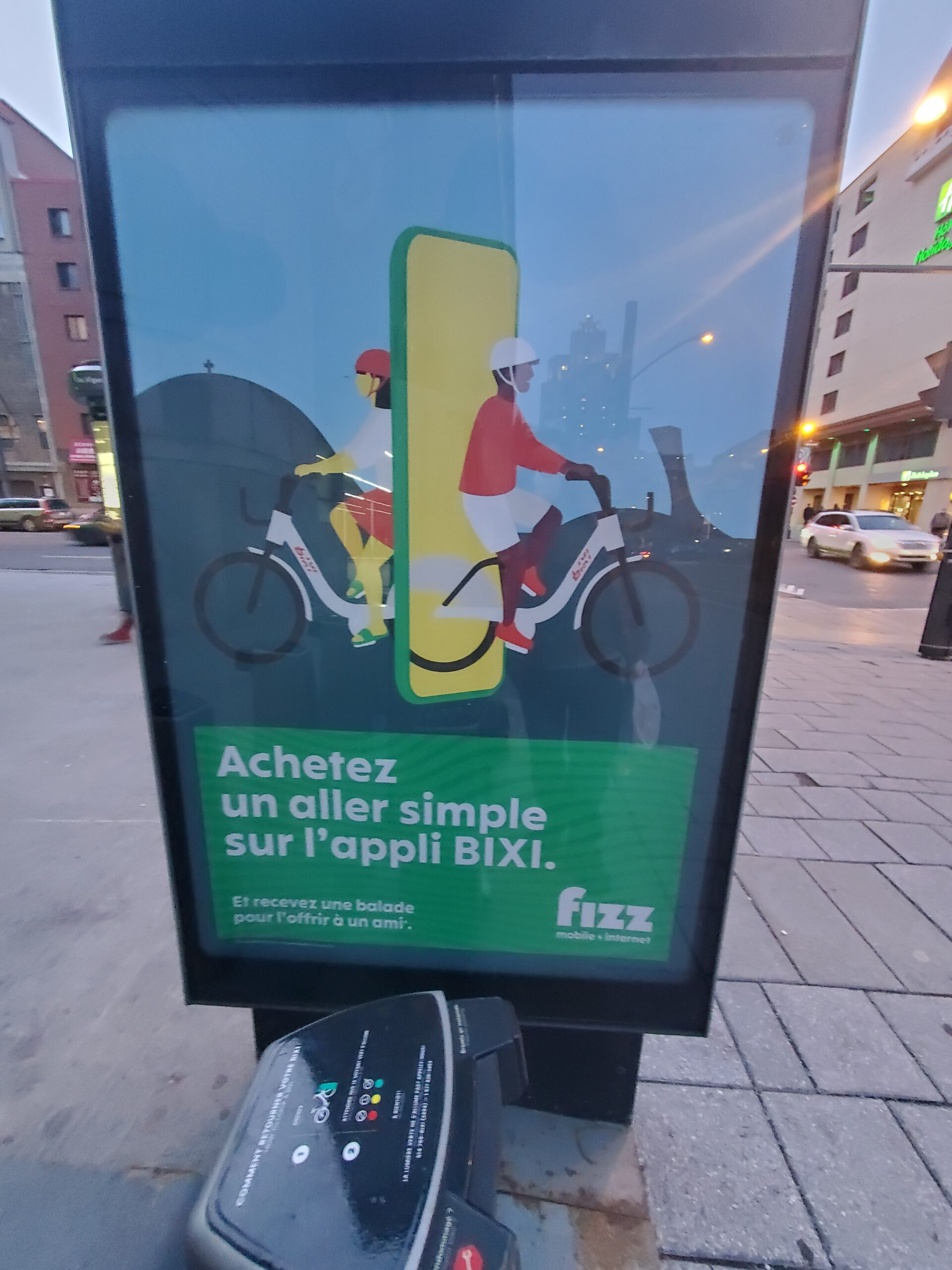 自行车停靠站一端的垂直广告牌。它展示了一款手机，手机两侧分别出现了背靠背的自行车。法国书上说，如果你买了一张Bixi单程票，你可以为你的朋友免费旅行。右下角写着Fizz Internet Mobile