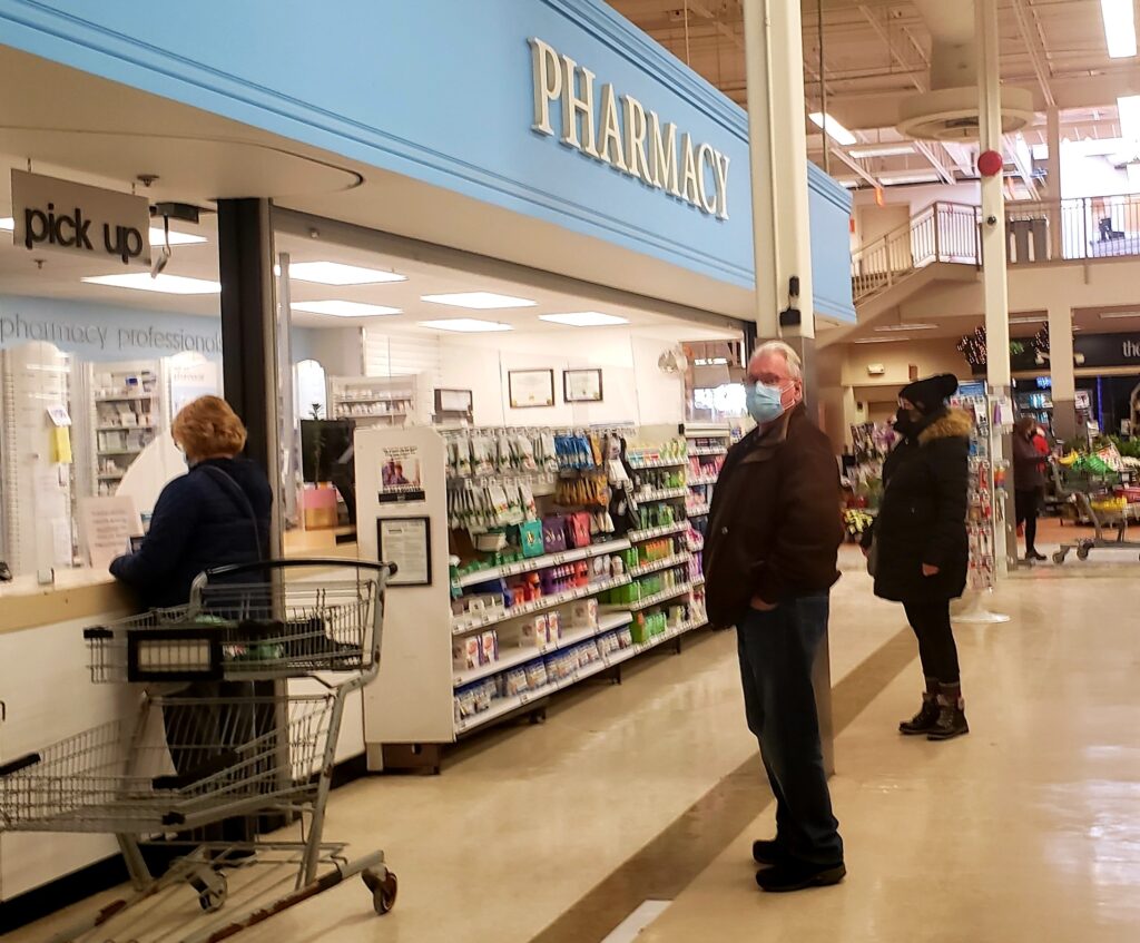 人们在下萨克维尔的超级商店的药房柜台前排队。柜台前面有一个架子，上面放着药品和其他产品。柜台上方是一块淡蓝色的牌子，上面写着“药房”。