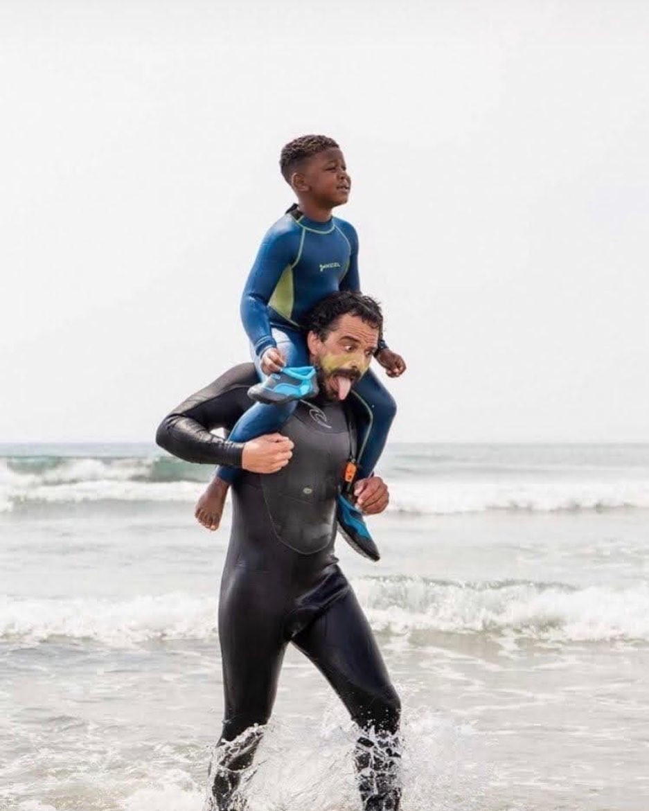 一个年轻的黑人男孩穿着一套湿漉漉的衣服，站在一个男人的肩膀上做鬼脸，他也穿着湿漉漉的衣服，站在白色天空的沙滩上。