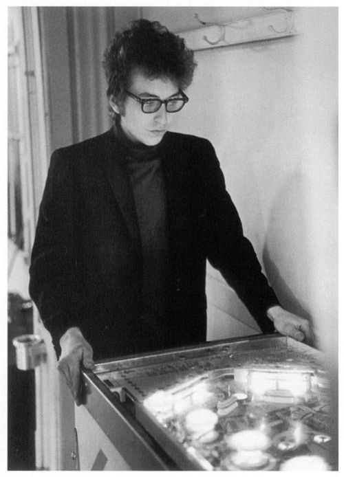 年轻的鲍勃·迪伦留着卷发，戴着眼镜，穿着深色夹克，双手放在弹球机的手柄上。