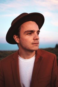 布莱登·林的照片，他是一个年轻的白人。他戴着一顶黑色的软呢帽，一件酒红色/铁锈色的西装外套，里面是一件白衬衫。