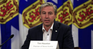 一个穿着深色西装、白衬衫、没有打领带的男人在讲台上发言。背景是四面新斯科舍旗，颜色分别为蓝色、黄色和红色。