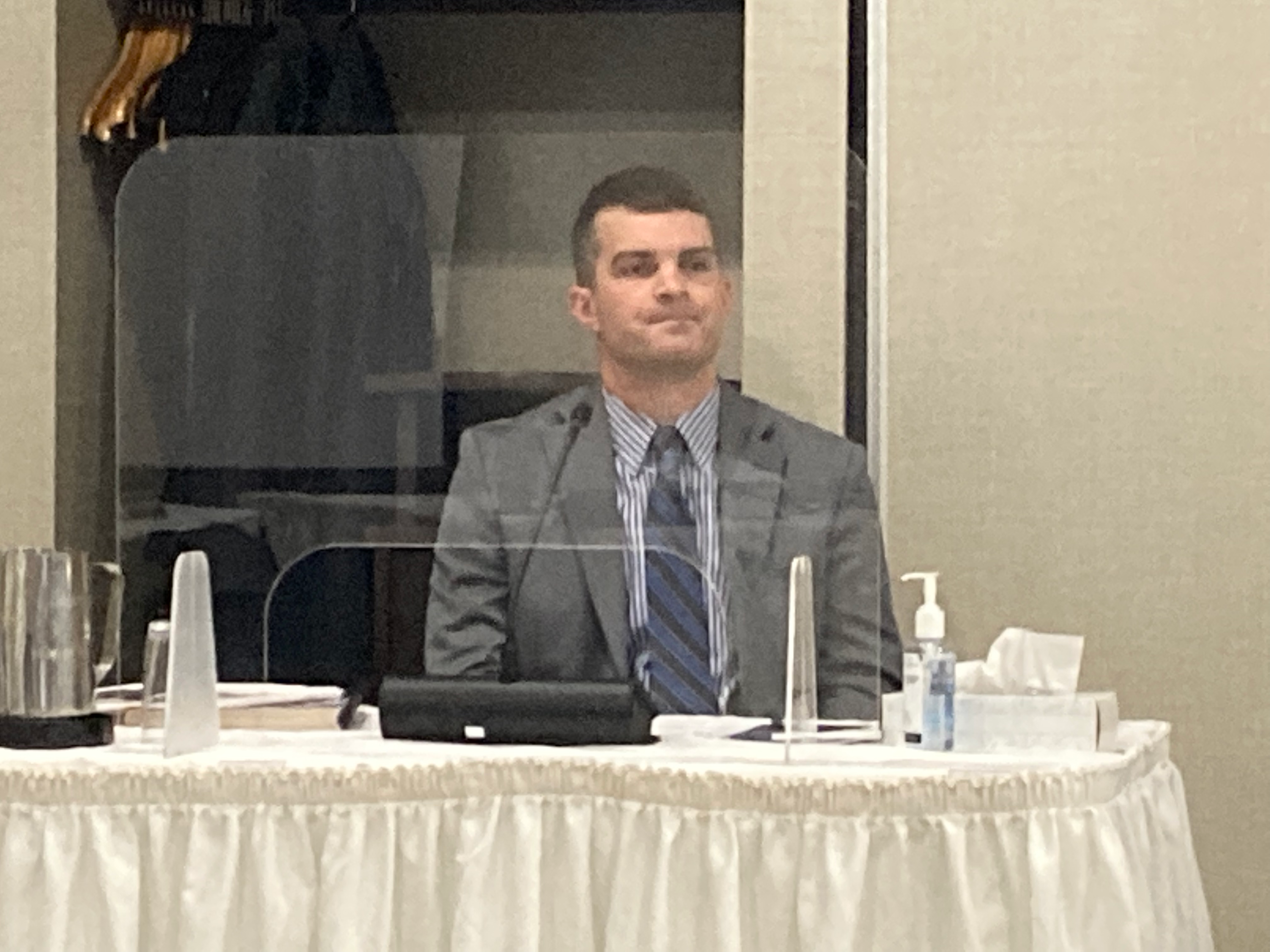 在证人席上，身穿灰色商务套间的白人男子坐在玻璃屏障后面