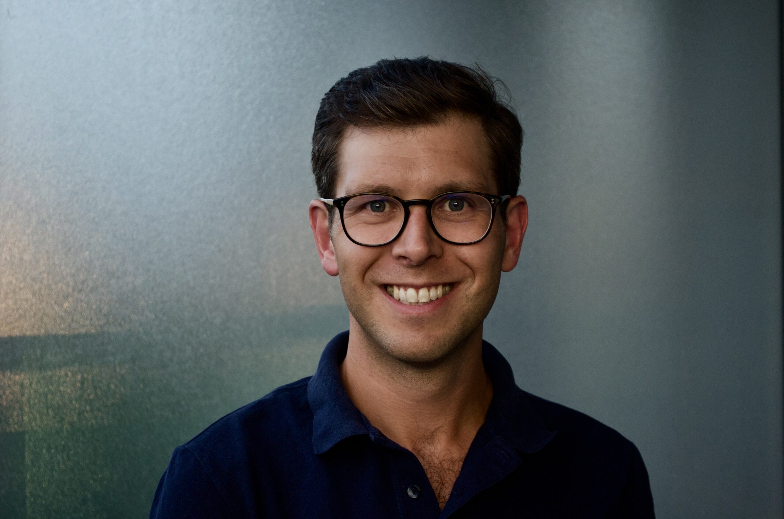 一名30多岁男子的头像，身穿深蓝色开领衬衫。他留着短发，从右边分开，戴着一副中性色的眼镜。他面带微笑，站在中性的蓝白色背景前。