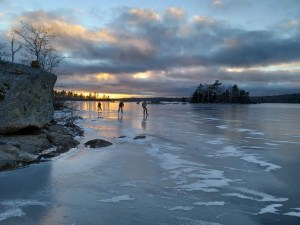 三个滑冰者经过结冰的加斯佩罗湖上的岩石和树木。背景是夕阳西下，金色的太阳映在地平线上