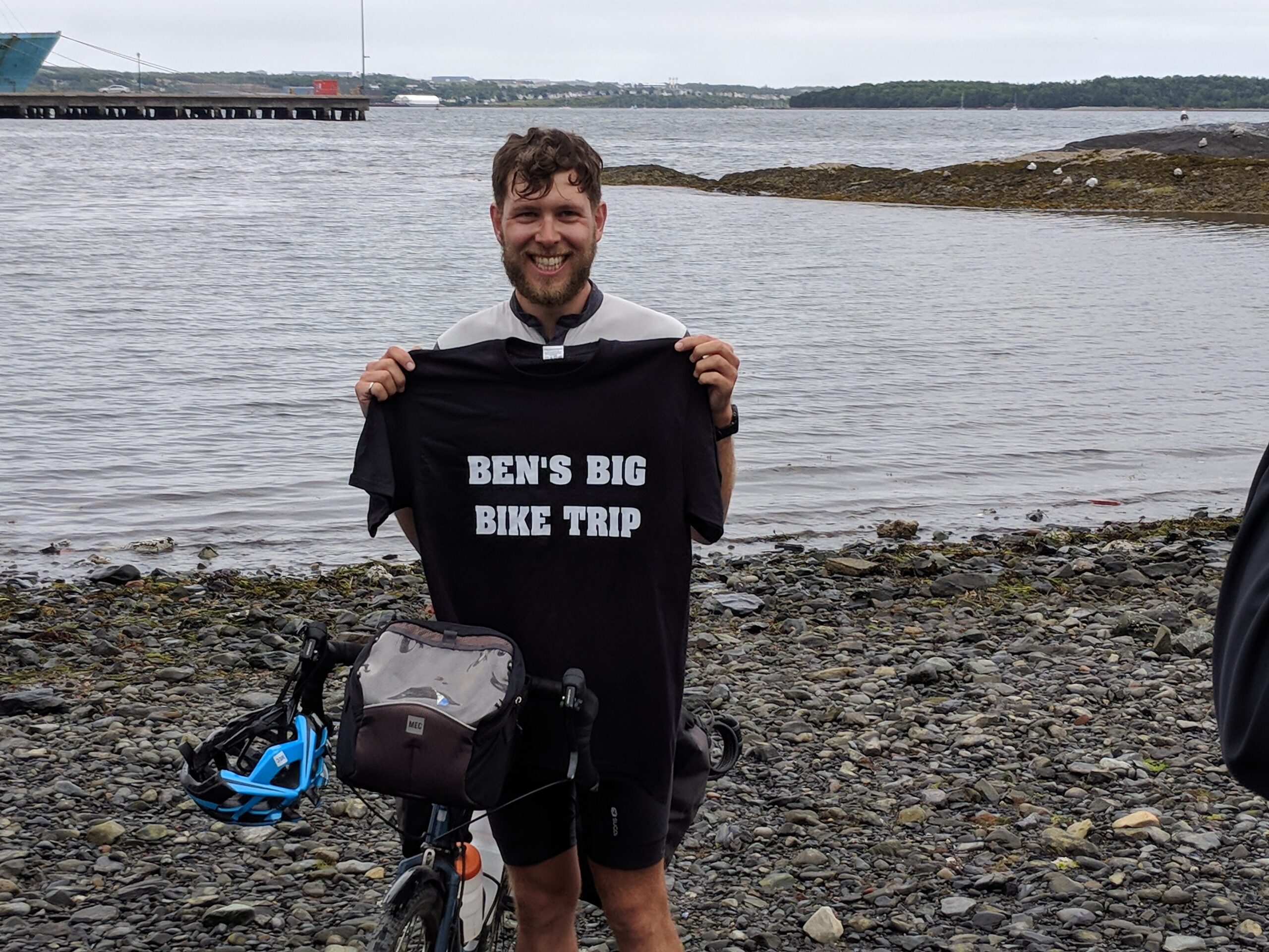 一个留着胡子的白人青年站在海边的岩石海滩上。他跨坐在自行车上，头盔挂在车把上。手里拿着一件写着“本的大自行车之旅”的白色t恤。