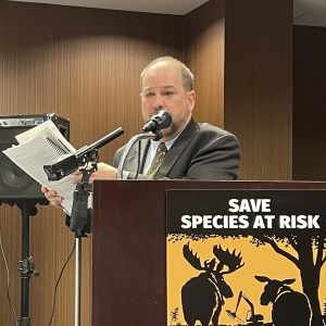Ray Plourde of EAC stands at a podium in a hotel conference room, speaking into a microphone, and holding a large pile of papers.