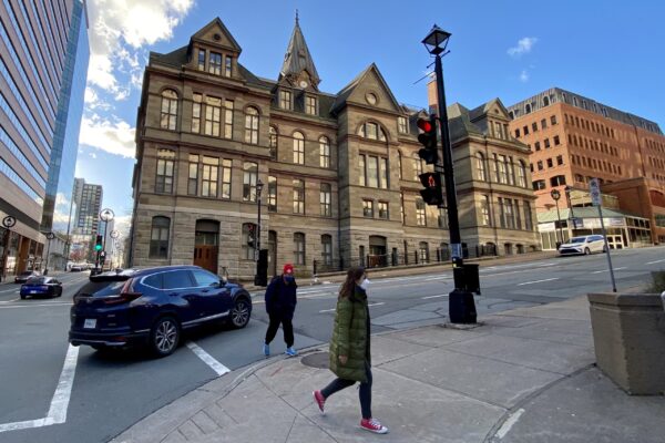 A city scene on a sunny winter day. In the background, the stone backside of Halifax City Hall. In the foreground, a pedestrian wearing a three-quarter length winter jacket, red sneakers and a white N95-style mask walks on the sidewalk. Another pedestrian behind her, wearing a red toque and blue sneakers, mounts the sidewalk. A right-turning navy SUV drives through a crosswalk behind the man.