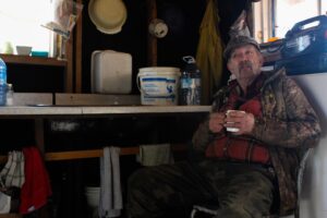 A middle aged, moustachioed man in outdoor camouflaged gear sits in the corner of his simple cabin, drinking tea.
