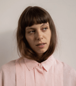 一张年轻的白人女子的照片，她留着棕色的长发和刘海。她的头发塞进浅粉红色的上衣里。