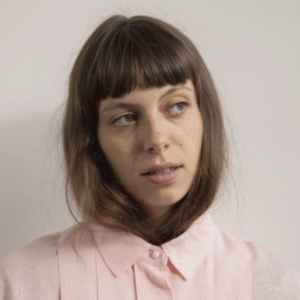 A photo of young white woman with long brown hair with bangs. Her hair is tucked into her pale pink blouse.