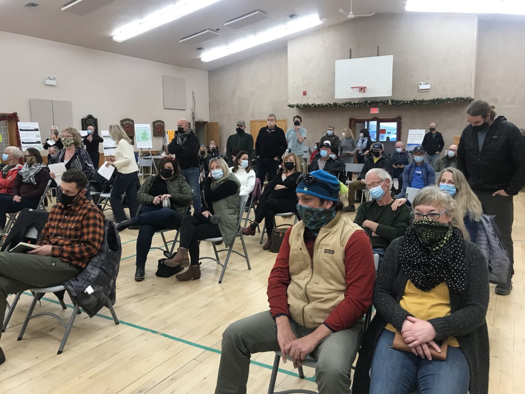 由于新冠疫情的间隔，温特沃斯娱乐中心的体育馆几乎挤满了数十人，他们参加了2021年12月4日在温特沃斯拟议的希金斯Mtn风电场的开放参观日，正如这张照片中所示，人们排队提问。图片:Joan Baxter