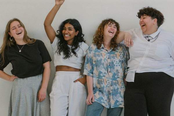 四个女人，三个白人，一个黑人，身材不一，站在白色的墙上笑着拍照。