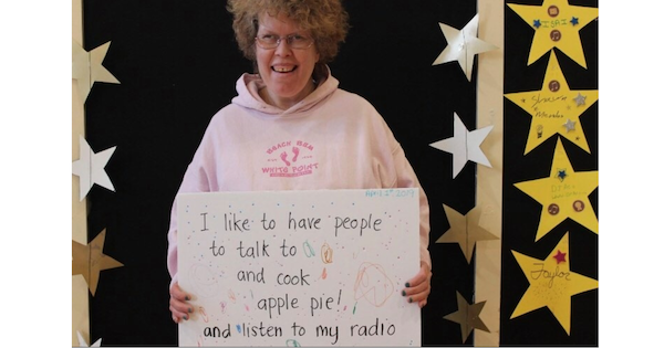 一个戴眼镜、穿粉色连帽衫的女人举着一个牌子，上面写着我喜欢有人带我去，有人做饭，有人吃苹果派，有人听我的收音机。