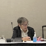 an older white woman with short grey hair and a dark suit jacket sits at a table with a microphone