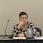 A white woman in a black and white plaid shirt sits at a table with a microphone