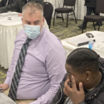 一个穿着薰衣草衬衫和条纹领带的白人，坐在桌子旁，有一个黑人妇女