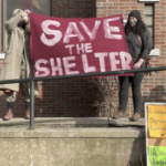 Two people on a set of steps at a brick building hold a red banner which reads 