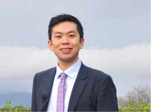 一名身穿灰色西装、白色衬衫、紫色领带的男子微笑着，背景是蓝色多云的天空和绿色植物。