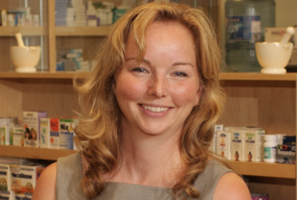 A smiling woman with reddish blond hair standing in front of a pharmacy counter with medication and a mortar and pestle in the background.