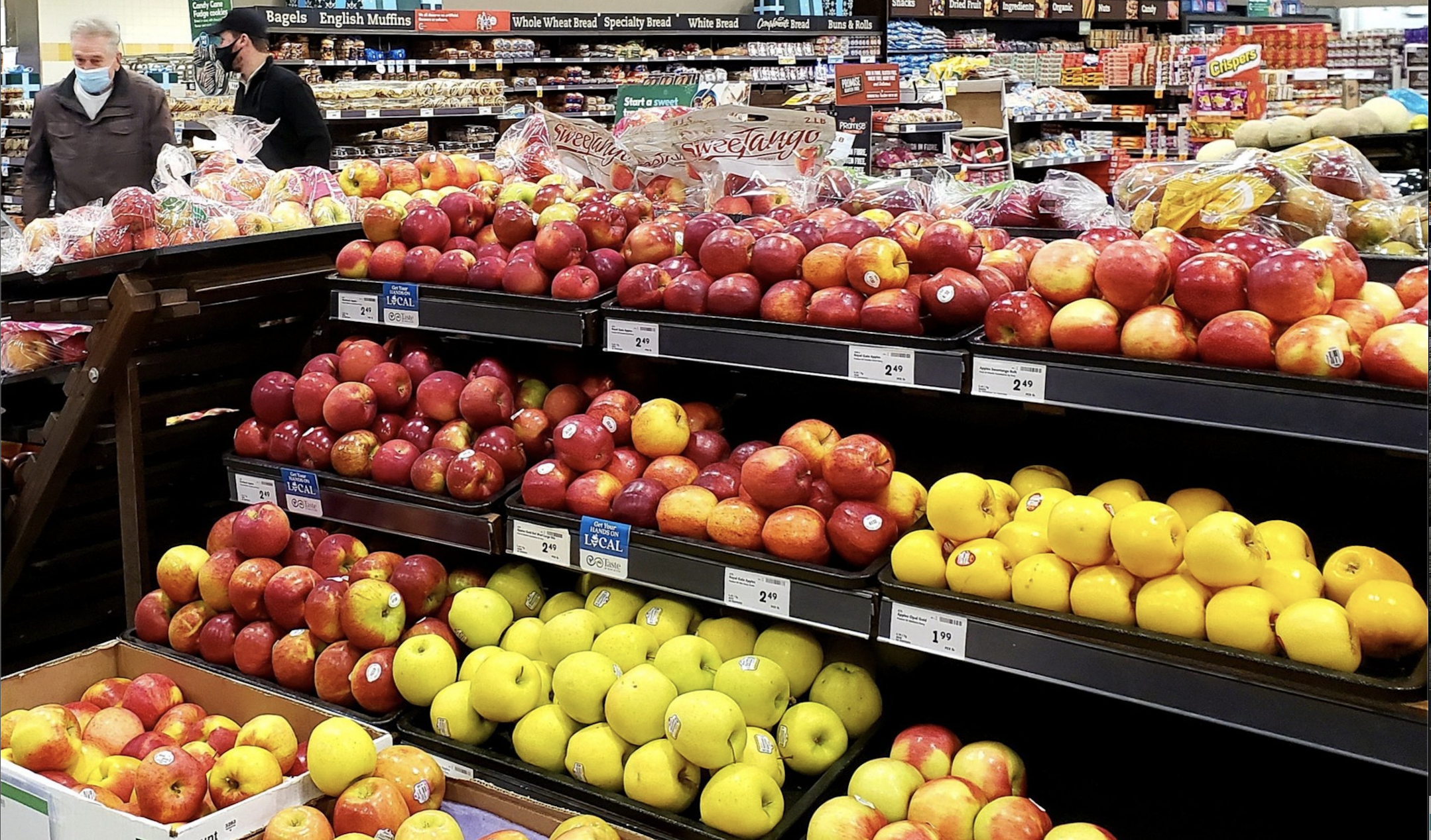 A grocery store display of various coloured apple varieties with a customer and grocery store worker in the background.