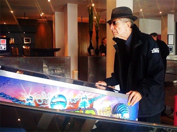 莱昂纳德·科恩身穿黑色夹克，头戴软呢帽，站在弹球机前