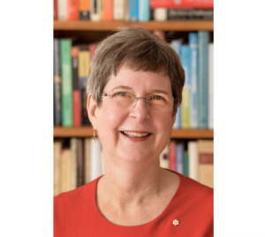 一个穿着鲜红色毛衣，戴着眼镜的女人对着镜头微笑，身后有一个书架。