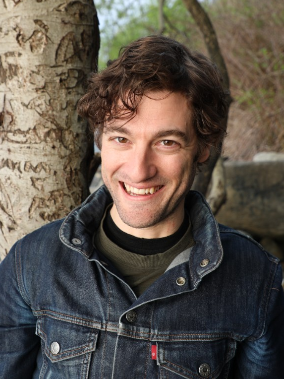 一个微笑的年轻白人男子的头像，穿着牛仔夹克，微微靠在树干上。